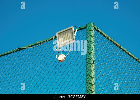 Projecteur LED monté sur la clôture verte de maillon de chaîne contre le ciel bleu. Mise au point sélective. Banque D'Images