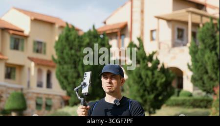Homme avec caméra à cardan se tient devant une villa de luxe dans un domaine ensoleillé Banque D'Images