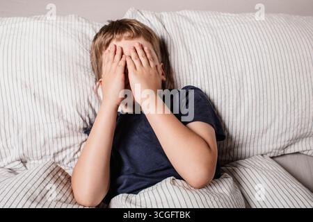 L'enfant avait peur avant d'aller au lit. Terreurs nocturnes chez un enfant. L'enfant couvre son visage avec ses mains dans une peur. Cauchemars et peur des ténèbres. Banque D'Images