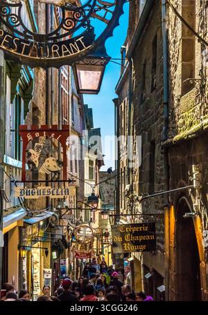 Vue rapprochée saisissante d’enseignes suspendues de style ancien se balançant au-dessus de la foule dans la rue principale du Mont-Saint-Michel, la Grande Rue. Beaucoup sont en fer forgé... Banque D'Images