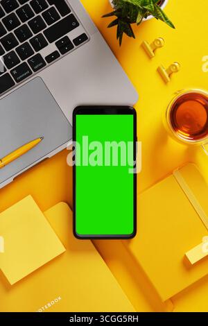 Smartphone avec écran vert près de l'ordinateur portable, stylo, cahiers, notes adhésives, plante, et du thé sur le bureau jaune. Présentez votre application, votre site Web ou votre capture d'écran. Banque D'Images