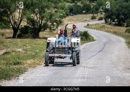 Un agriculteur âgé conduit avec sa femme assis sur le garde-boue de leur vieux tracteur bleu des champs le long d'une piste dans la Turquie rurale Banque D'Images