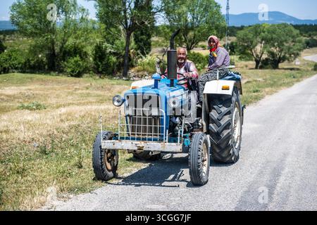 Un agriculteur âgé conduit avec sa femme assis sur le garde-boue de leur vieux tracteur bleu des champs le long d'une piste dans la Turquie rurale Banque D'Images