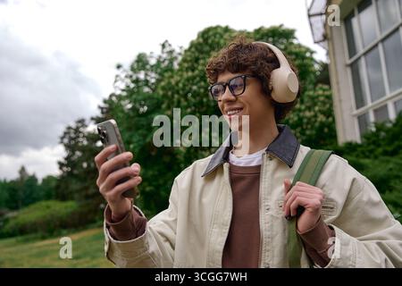 Un jeune homme se promène à l'extérieur, souriant en se connectant avec des amis via son smartphone. Banque D'Images