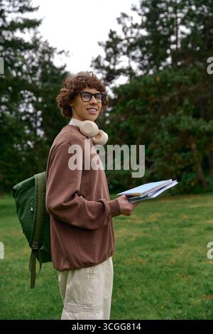 Un jeune homme beau aux cheveux bouclés marche dehors, tenant des livres et portant des écouteurs dans la nature. Banque D'Images