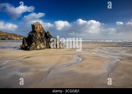 Formation rocheuse se dresse sur une plage de sable, entourée de marées d'eau sous un ciel bleu profond avec des nuages dispersés et des vagues au loin Banque D'Images