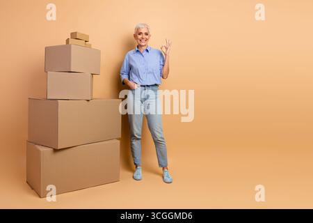 Femme senior dans la tenue élégante gestuelle positivement avec des boîtes en carton sur fond Beige minimal Banque D'Images