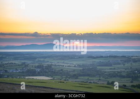 Un coucher de soleil chaud descend derrière des collines lointaines, projetant des teintes dorées et violettes sur de vastes terres agricoles et des champs en patchwork. Les moutons paissent paisiblement Banque D'Images
