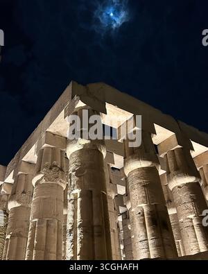 Louxor, Égypte - 9 février 2025 : vue nocturne des piliers illuminés du Temple de Louxor avec la lune dans le ciel partiellement enveloppée par des nuages à Louxor, Égypte. Banque D'Images
