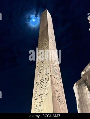 Louxor, Égypte - 9 février 2025 : vue nocturne de l'obélisque illuminé du pharaon Ramsès II au Temple de Louxor en Égypte, avec la lune brillante à travers les nuages Banque D'Images