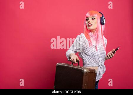 Jeune touriste tenant une valise et un smartphone, portant des écouteurs sans fil, appréciant la musique et cherchant sa porte d'embarquement. Jolie femme caucasienne prête à voyager avec des bagages. Banque D'Images