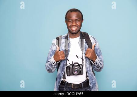 Portrait d'un jeune homme noir joyeux donnant deux pouces vers le haut, avec son reflex numérique et sac à dos sur lui. Heureux photographe masculin regardant l'appareil photo, faisant un geste d'accord avec ses pouces. Banque D'Images