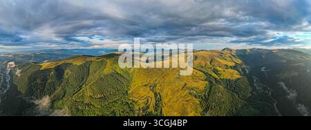 Vue panoramique aérienne des montagnes alpines avec des vallées boisées verdoyantes, des routes sinueuses et des crêtes herbeuses sous un ciel nuageux spectaculaire en été Banque D'Images
