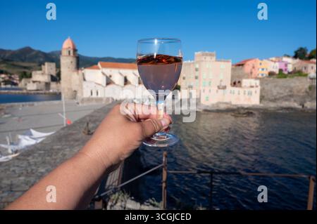 Main avec verre de vin rose Collioure AOC aux rayons du soleil avec vue sur la vieille ville de Collioure, maisons colorées, église, château et plages, Occitanie, franc Banque D'Images