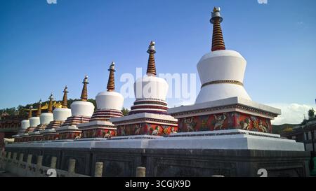 De nombreux stupas blancs se dressent dans une rangée sous un ciel clair, mettant en valeur leurs structures en couches et leurs bases complexes, avec un soupçon de montagnes dans le backgroun Banque D'Images