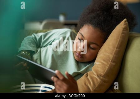 Enfant noir couché sur le canapé à l'aide d'une tablette numérique, reposant la tête sur le coussin tout en se concentrant sur l'écran, tenant le dispositif avec les deux mains, posture détendue, cadre intérieur décontracté Banque D'Images