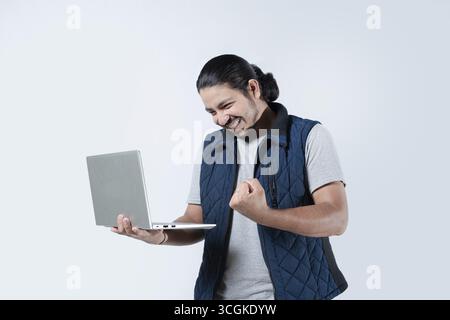 Homme célébrant avec le poing dans l'air tenant un ordinateur portable sur fond blanc. Jeune homme célébrant tenant un ordinateur sur fond isolé Banque D'Images