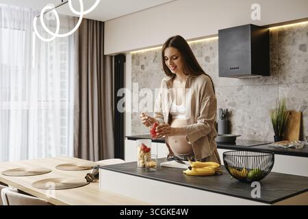 Une femme enceinte prépare joyeusement une collation aux fruits saine dans sa cuisine élégante Banque D'Images