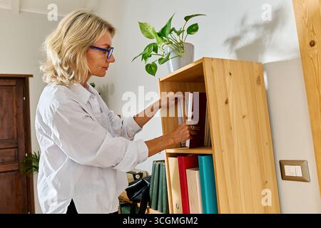 Femme d'affaires aux cheveux blonds cherchant des informations et prenant un livre de bibliothèque Banque D'Images