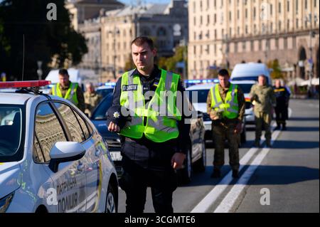 Kiev, Ukraine - 29 août 2025 : policier ukrainien tenant son chapeau à côté d'une voiture de patrouille, entouré d'officiers et de soldats en formation dans une rue de la ville lors de l'hommage aux héros tombés au combat lors de la Journée du souvenir des défenseurs de l'Ukraine Banque D'Images