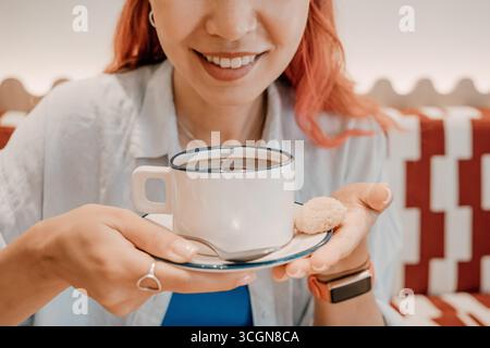 Femme aux cheveux roses vibrants souriant tout en tenant une tasse de café blanc, appréciant un petit biscuit placé sur une soucoupe Banque D'Images
