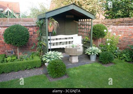 Un charmant coin salon extérieur niché dans un jardin, avec un banc en bois, des plantes décoratives et une table en pierre, rayonnant de tranquillité. Banque D'Images