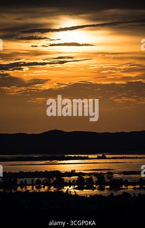 Fleuve Ayeyarwaddy Couchant doré depuis la colline de Mandalay Mandalay Myanmar | MANDALAY, Myanmar — Une bande lumineuse de lumière du coucher de soleil marque le chemin du fleuve Ayeyarwaddy à travers les plaines en contrebas de la colline de Mandalay. La colline, un site sacré s'élevant à 754 pieds (230 m), surplombe un système fluvial drainant environ 411 000 à 421 000 kilomètres carrés, soit environ deux tiers de la superficie du Myanmar. Mandalay, l'ancienne capitale royale de la dynastie Konbaung, reste un centre économique et culturel majeur au centre du Myanmar. Banque D'Images