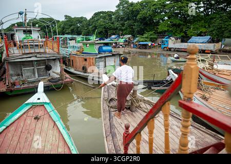 Bateaux en bois de la rivière Ayeyarwady amarrés à la rivière Port Mandalay Myanmar // MANDALAY, Myanmar — Une femme sur un bateau traditionnel en bois gère les cordes alors qu'il accoste à un port fluvial animé sur la rivière Ayeyarwady (également connue sous le nom de rivière Irrawaddy). De nombreux autres bateaux en bois, essentiels pour le transport de marchandises et de passagers, sont amarrés le long de la rive boueuse. La rivière Ayeyarwady est la voie navigable commerciale la plus longue et la plus cruciale du Myanmar, facilitant le commerce et les déplacements à travers le pays. Ce port actif de Mandalay, la deuxième plus grande ville et un centre économique important de la région de Mandalay, est une hu Banque D'Images