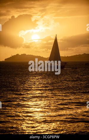 Voilier sur l'océan au coucher du soleil St Thomas de St John USVI // ST JOHN, USVI — Un voilier navigue sur les eaux au large de John lorsque le soleil se couche, projetant une lueur dorée sur l'horizon. Au loin, l’île de produits Thomas est visible, silhouettée contre le ciel vibrant. La vue est capturée depuis Gallows point dans Cruz Bay, un point de vue populaire sur le lieu John. Les îles Vierges américaines sont un groupe d'îles de la mer des Caraïbes, au sud-est de Porto Rico. Banque D'Images
