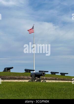 Rangée de canons historiques positionnés le long des remparts herbeux du Fort Macon State Park en Caroline du Nord, avec le drapeau américain flottant fièrement au-dessus. Banque D'Images