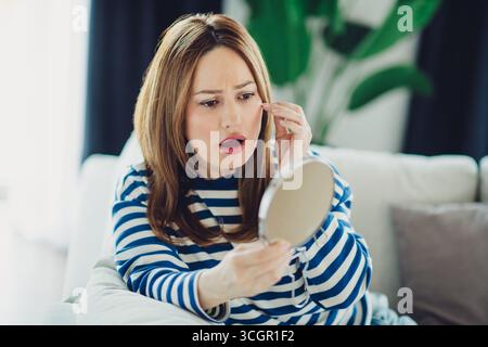 Jeune femme en tenue décontractée rayée admirant son reflet dans un miroir de maquillage à la maison pendant une journée détendue Banque D'Images