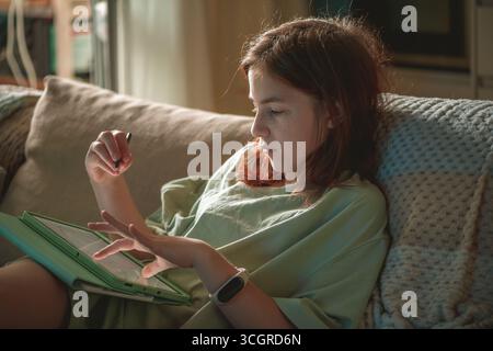 Se concentrant sur son art numérique, une jeune fille préadolescente dans des vêtements décontractés dessine sur une tablette avec un stylet, profitant du temps libre tout en se relaxant sur un sof confortable Banque D'Images