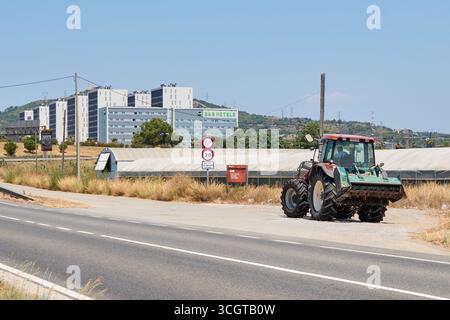 Viladecans. Barcelone - 29 août 2025 : un tracteur agricole stationné sur l'épaule, avec des serres et un complexe hôtelier et d'appartements B moderne Banque D'Images