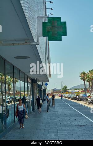 Viladecans. Barcelone - 29 août 2025 : une femme marche vers la caméra sur un large trottoir à Viladecans, sous la croix verte d'une pharmacie. Banque D'Images