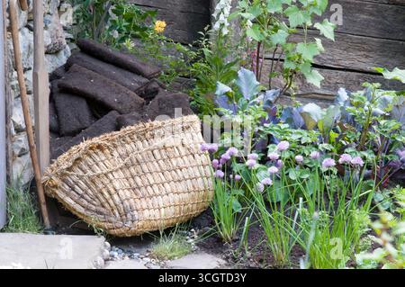 Un panier tissé fabriqué à la main s'appuie contre une clôture en bois dans un jardin luxuriant rempli de fleurs colorées, d'herbes et de terre. Banque D'Images