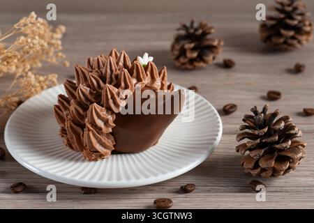 Gros plan d'un dessert au chocolat en forme de hérisson sur une assiette blanche, stylisé avec des pommes de pin, des grains de café et des fleurs séchées pour une sensation confortable et rustique Banque D'Images