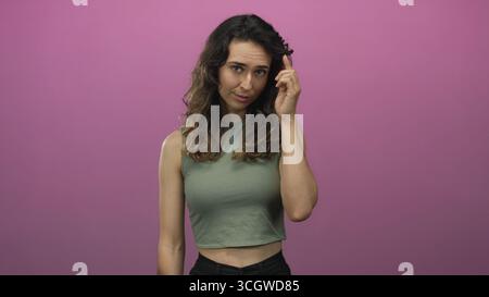 Femme hispanique dans le top vert de la récolte avec les cheveux ondulés et l'expression réfléchie pointant le doigt vers le temple dans le studio ; contemplation pensée réflexion idée. Banque D'Images