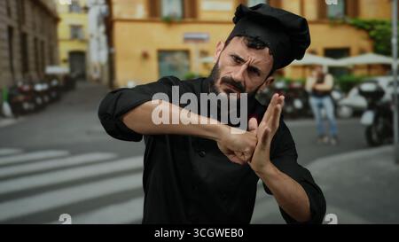Homme hispanique barbu habillé comme chef imitant la pose de boxe dans une rue extérieure, mêlant tenue professionnelle et expression ludique. Banque D'Images
