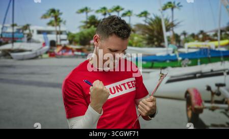 Homme portant une chemise rouge de volontaire debout sur le front de mer tenant une planche à pince montrant des pouces vers le haut expression confiante au milieu des palmiers et des bateaux suggérant outd Banque D'Images