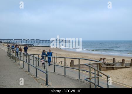 Les gens marchent le long de la promenade par la plage de Southwold, avec Southwold Pier s'étendant dans la mer du Nord en arrière-plan, Suffolk, Angleterre. Banque D'Images