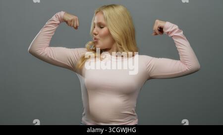 Femme fléchissant les muscles en toute confiance sur un fond gris lisse, portant une chemise rose décontractée, incarnant la force, l'autonomisation, la forme physique et la positiv Banque D'Images