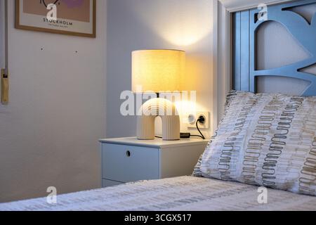Vue latérale d'un coin chambre avec une lampe de table crème sur une table de chevet à côté d'un lit soigneusement fait. Banque D'Images