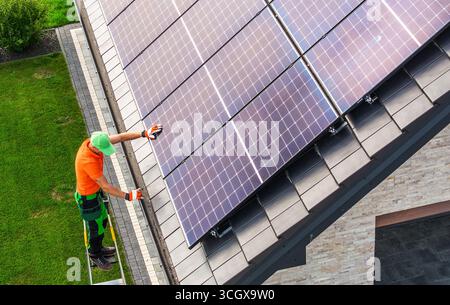 Un technicien portant un équipement de sécurité installe des panneaux solaires sur un toit résidentiel pendant la journée, mettant en valeur les efforts en matière d’énergie verte et la technologie moderne. Banque D'Images