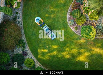Une personne tond une pelouse verte luxuriante dans un jardin magnifiquement aménagé avec des parterres de fleurs et des plantes décoratives pendant la lumière du jour. Banque D'Images