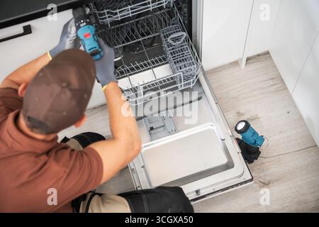 Un technicien travaille sur la réparation d'un lave-vaisselle dans une cuisine moderne, à l'aide d'un tournevis et en examinant attentivement l'appareil. Banque D'Images