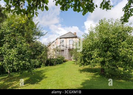 Une vieille maison abandonnée en bois avec une façade usée et un petit porche est entourée d'arbres et d'une pelouse verte par une journée ensoleillée. Banque D'Images