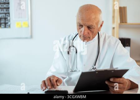 Docteur comptable masculin senior âgé en manteau blanc avec stéthoscope calculant les finances de comptabilité et le budget de l'hôpital à l'intérieur de la clinique, concept o Banque D'Images