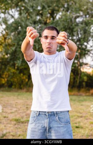 Jeune homme montrant un geste du pouce vers le bas dans un parc exprimant sa désapprobation Banque D'Images