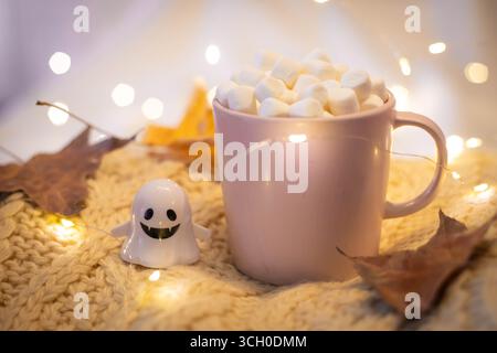 Halloween nature morte avec tasse rose remplie de chocolat et de guimauves, petit fantôme et guirlande lumineuse sur un pull en laine blanche. Festif, dos confortable Banque D'Images