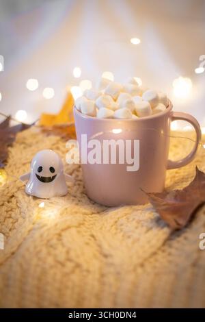 Halloween nature morte avec tasse rose remplie de chocolat et de guimauves, petit fantôme et guirlande lumineuse sur un pull en laine blanche. Festif, dos confortable Banque D'Images